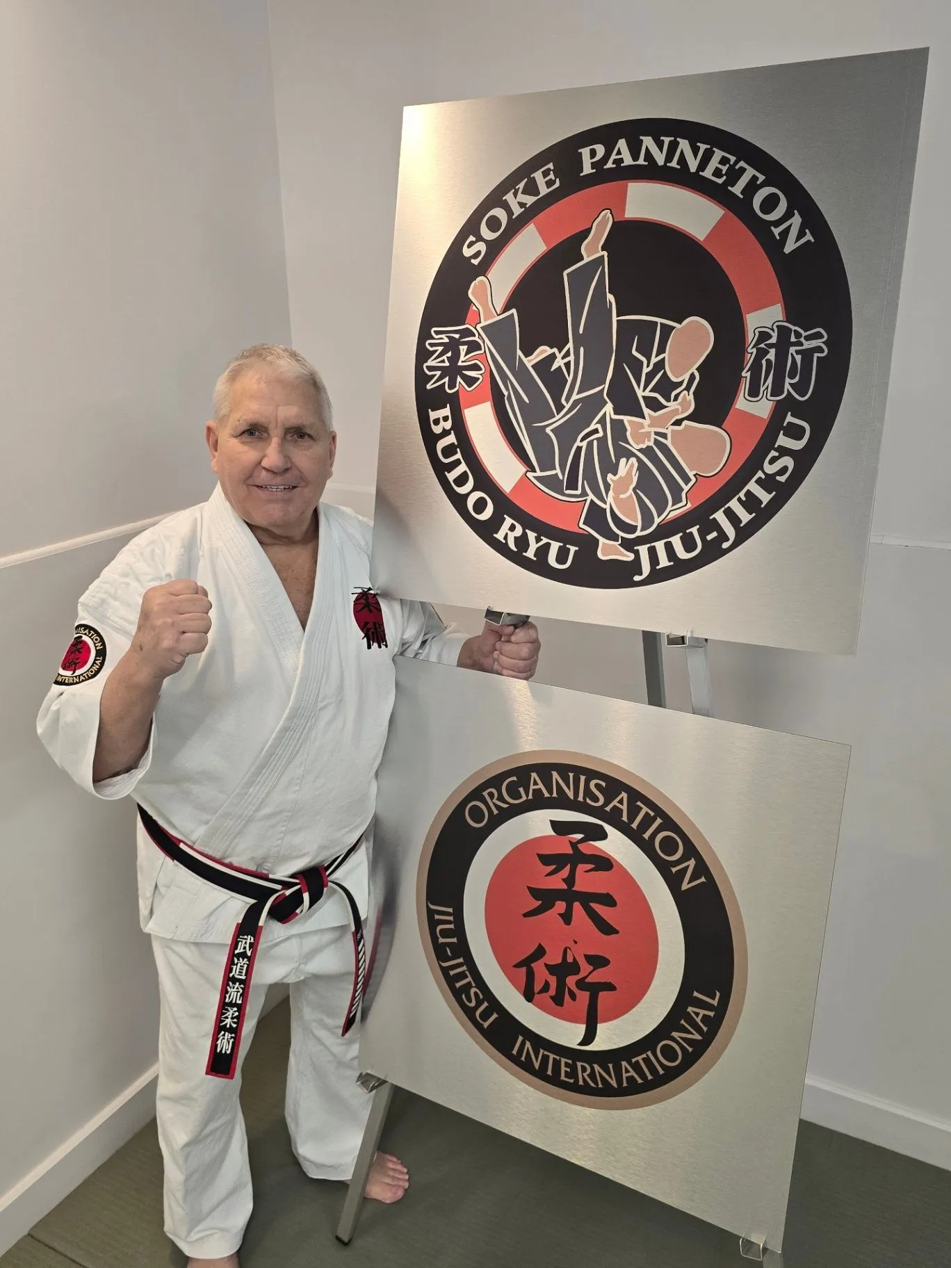 Soke Robert Panneton en kimono blanc près des écussons du Budo-Ryu et de l’OJI